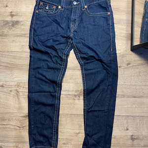 May include: Dark blue denim jeans with orange stitching. The jeans have a button and zipper closure, front and back pockets, and a classic five-pocket design. The jeans are laid flat on a wooden surface.