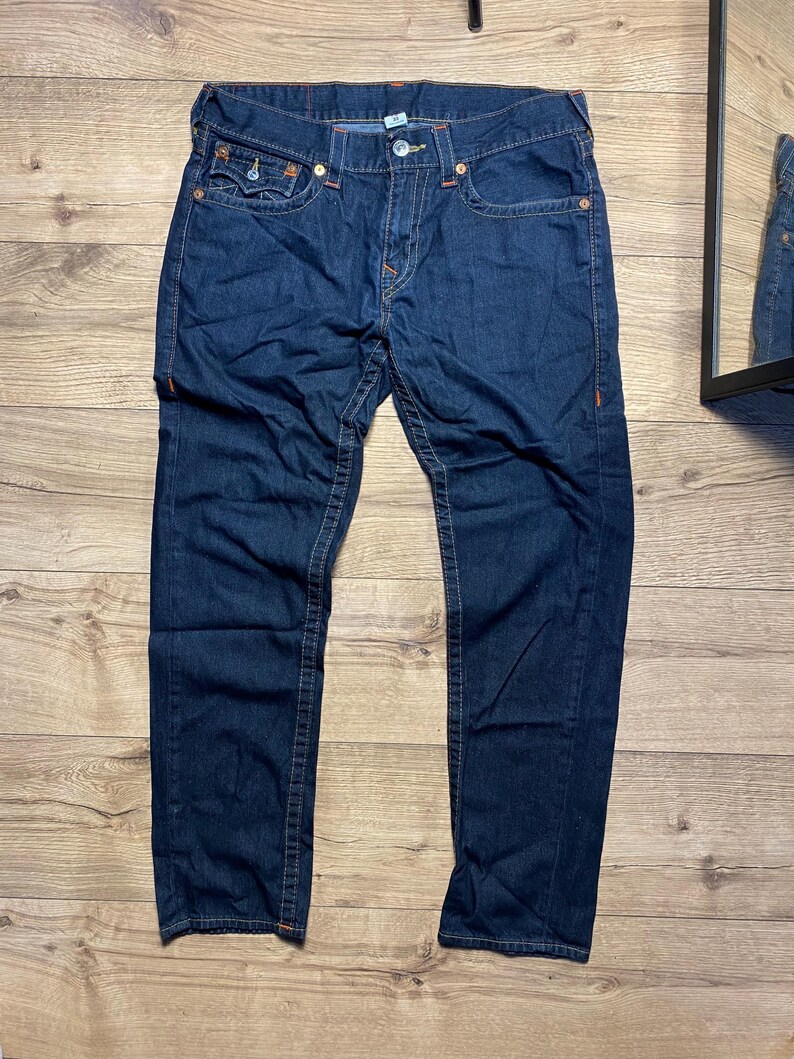May include: Dark blue denim jeans with orange stitching. The jeans have a button and zipper closure, front and back pockets, and a classic five-pocket design. The jeans are laid flat on a wooden surface.