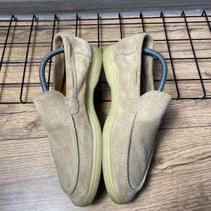 Op de afbeelding: Een paar beige su&egrave;de loafers met een gestikte neus in mocassin-stijl, een laag profiel en een lichtgekleurde rubberen zool. Elke schoen bevat een zilverkleurige metalen schoenspanner om de vorm van het schoeisel te behouden.