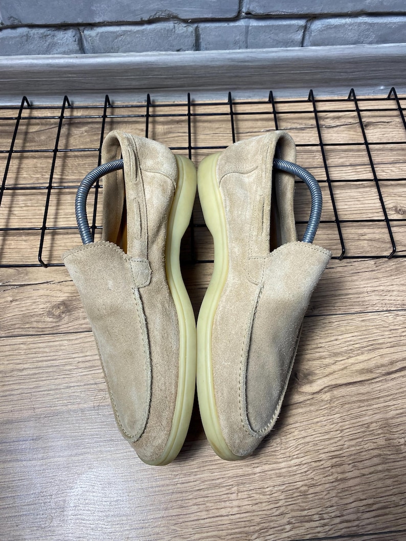 Op de afbeelding: Een paar beige su&egrave;de loafers met een gestikte neus in mocassin-stijl, een laag profiel en een lichtgekleurde rubberen zool. Elke schoen bevat een zilverkleurige metalen schoenspanner om de vorm van het schoeisel te behouden.