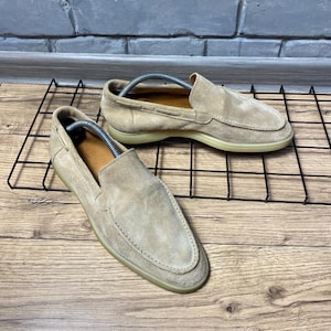 Op de afbeelding: Een paar beige su&egrave;de loafers met een gestikte neus in mocassin-stijl, een laag profiel en een platte rubberen zool. Elke schoen bevat een zilverkleurige metalen schoenspanner met veer om de vorm van het schoeisel te behouden.