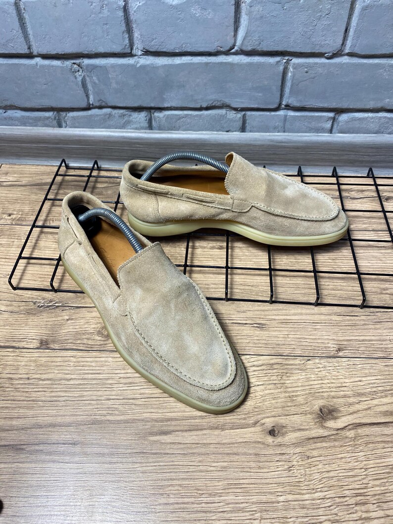 Op de afbeelding: Een paar beige su&egrave;de loafers met een gestikte neus in mocassin-stijl, een laag profiel en een platte rubberen zool. Elke schoen bevat een zilverkleurige metalen schoenspanner met veer om de vorm van het schoeisel te behouden.