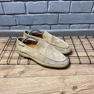 Op de afbeelding: Een paar beige su&egrave;de instappers met een mocassin-stijl gestikte neus, een laag profiel en een platte, lichtgekleurde rubberen zool. Elke schoen bevat een zilverkleurige metalen schoenspanner om de vorm van de schoen te behouden.