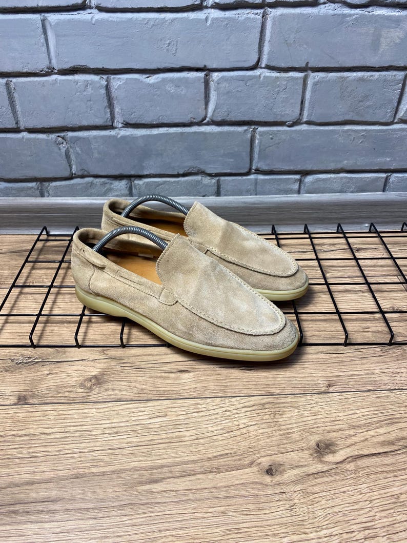 Op de afbeelding: Een paar beige su&egrave;de instappers met een mocassin-stijl gestikte neus, een laag profiel en een platte, lichtgekleurde rubberen zool. Elke schoen bevat een zilverkleurige metalen schoenspanner om de vorm van de schoen te behouden.