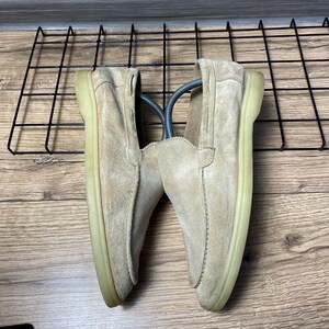 Op de afbeelding: Een paar beige su&egrave;de loafers met een mocassin-stijl gestikte neusnaad, een laag profiel en een doorschijnende rubberen zool. De schoenen zijn voorzien van zilverkleurige metalen schoenspanners om hun vorm te behouden.