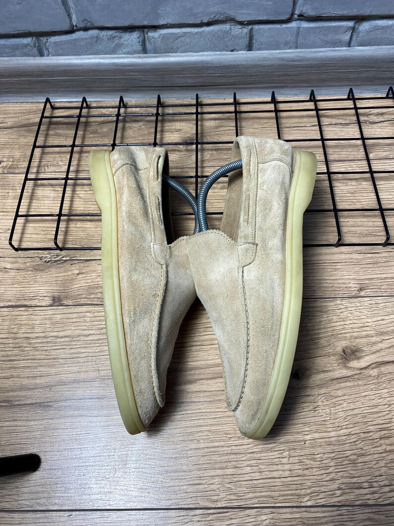 Op de afbeelding: Een paar beige su&egrave;de loafers met een mocassin-stijl gestikte neusnaad, een laag profiel en een doorschijnende rubberen zool. De schoenen zijn voorzien van zilverkleurige metalen schoenspanners om hun vorm te behouden.
