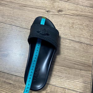 May include: A black slide sandal featuring a Medusa head emblem and a Greek key pattern on the strap. The word VERSACE and Made in Italy are embossed on the footbed. A blue measuring tape is placed over the sandal, showing a length of 26 cm.