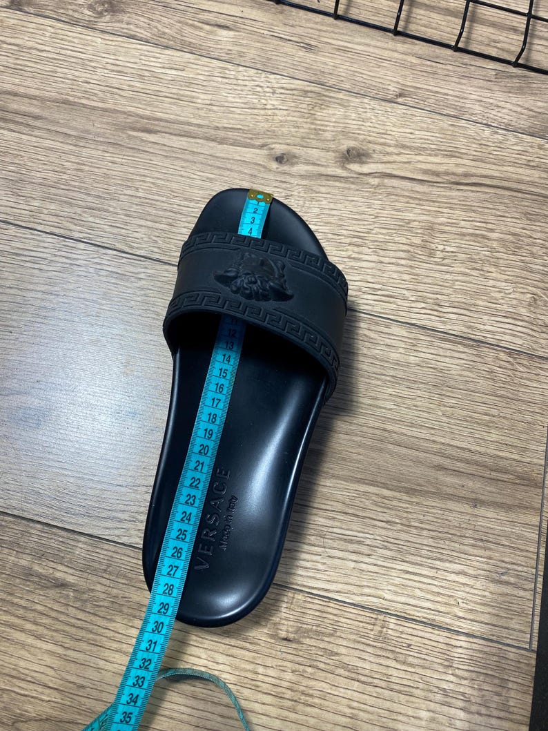 May include: A black slide sandal featuring a Medusa head emblem and a Greek key pattern on the strap. The word VERSACE and Made in Italy are embossed on the footbed. A blue measuring tape is placed over the sandal, showing a length of 26 cm.