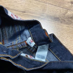 May include: Close-up of dark blue denim jeans with orange stitching. The jeans have a size 33 tag and a label with text in multiple languages, including "MADE IN MEXICO" and "100% COTTON". The jeans are folded, showing the inside waistband.