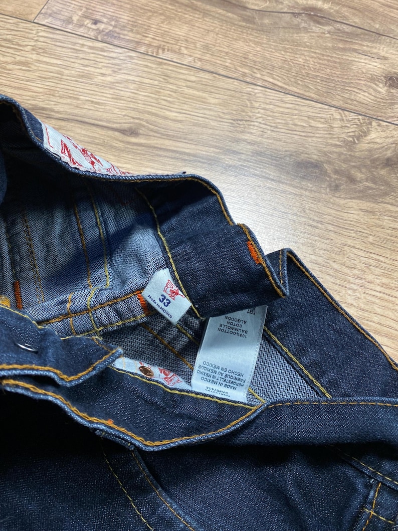May include: Close-up of dark blue denim jeans with orange stitching. The jeans have a size 33 tag and a label with text in multiple languages, including "MADE IN MEXICO" and "100% COTTON". The jeans are folded, showing the inside waistband.