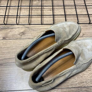 Op de afbeelding: Een paar beige su&egrave;de loafers met een mocassin-stijl neus, een laag profiel en bruinleren inlegzolen met de merknaam Aurelien in schrijfletters gedrukt. Elke schoen is voorzien van een zilverkleurige metalen veerspanner voor schoenen.