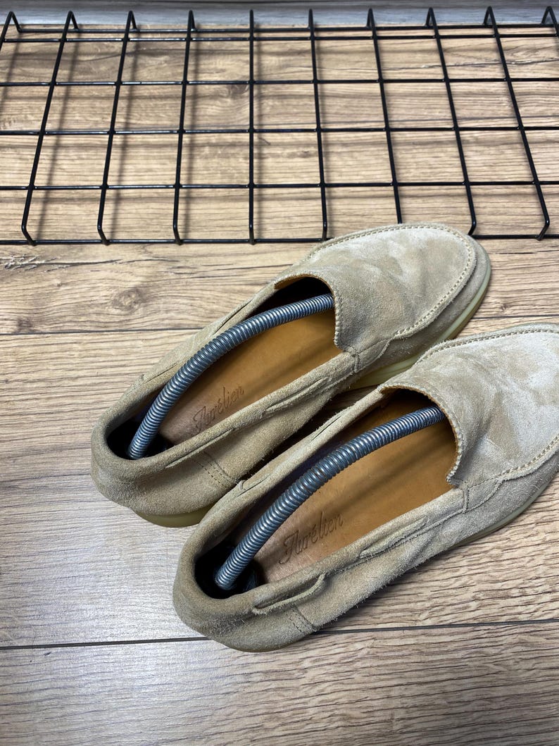 Op de afbeelding: Een paar beige su&egrave;de loafers met een mocassin-stijl neus, een laag profiel en bruinleren inlegzolen met de merknaam Aurelien in schrijfletters gedrukt. Elke schoen is voorzien van een zilverkleurige metalen veerspanner voor schoenen.