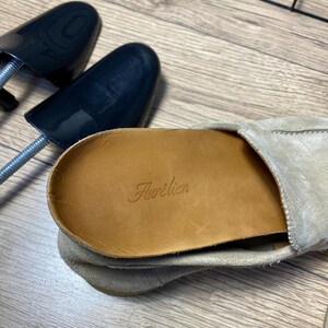 Op de afbeelding: Een beige su&egrave;de loafer met een lichtbruine leren binnenzool waarop de merknaam Aur&eacute;lien in reli&euml;f is aangebracht. Een zwarte plastic schoenspanner ligt naast de schoen, die zichtbare stiksels aan de zijkant en een zachte, getextureerde afwerking heeft.