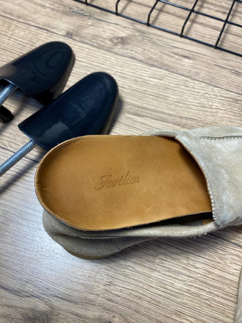 Op de afbeelding: Een beige su&egrave;de loafer met een lichtbruine leren binnenzool waarop de merknaam Aur&eacute;lien in reli&euml;f is aangebracht. Een zwarte plastic schoenspanner ligt naast de schoen, die zichtbare stiksels aan de zijkant en een zachte, getextureerde afwerking heeft.