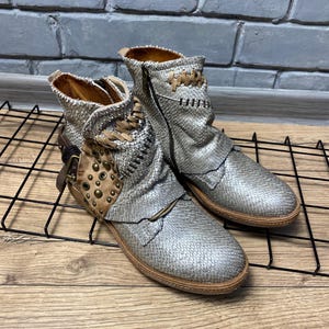 May include: A pair of silver ankle boots with a textured snakeskin-like pattern. The boots feature tan leather side panels adorned with metallic studs, a side buckle strap, and decorative tan stitching details along the collar and upper shaft.