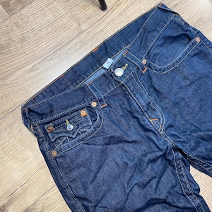 May include: Dark blue denim jeans with orange stitching and metal rivets. The jeans have a classic five-pocket design and a button and zipper closure. The jeans are size 32.