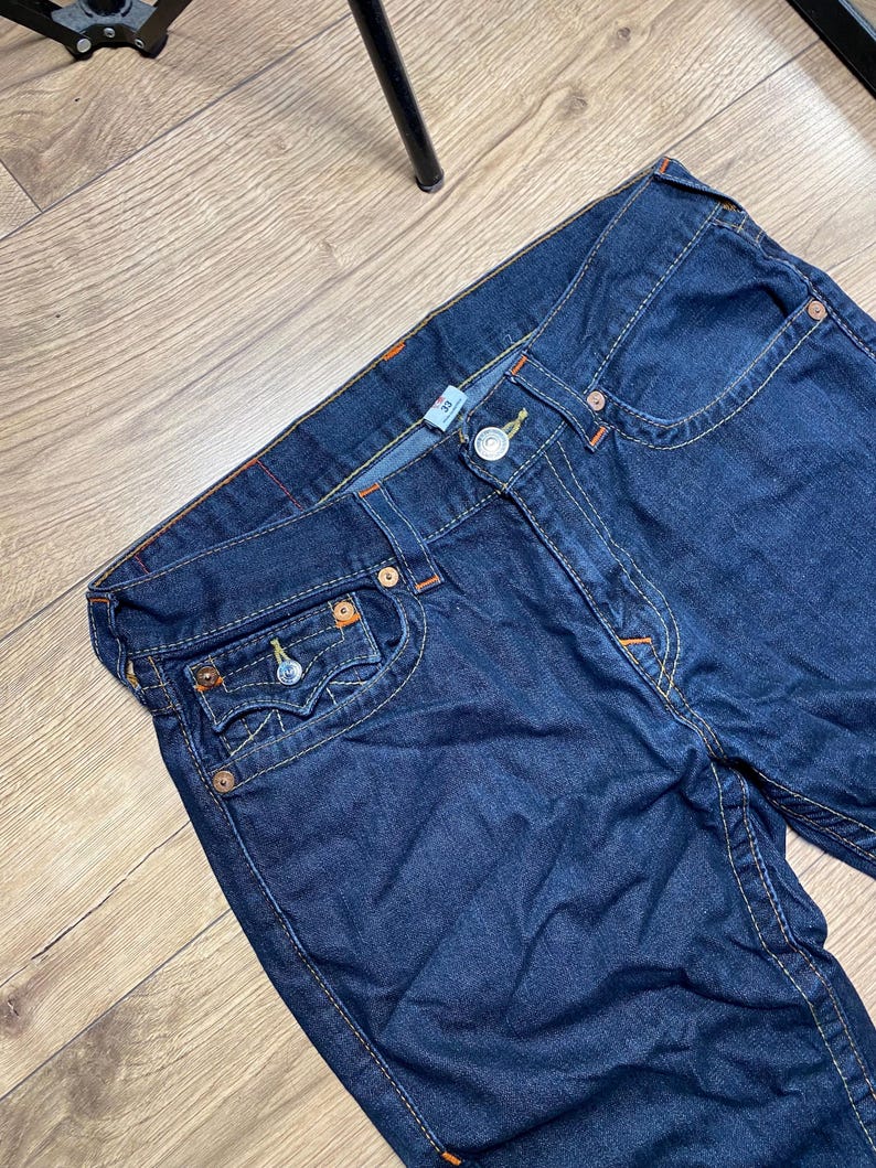 May include: Dark blue denim jeans with orange stitching and metal rivets. The jeans have a classic five-pocket design and a button and zipper closure. The jeans are size 32.