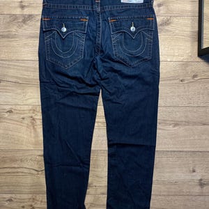 May include: Dark blue denim jeans with white stitching and orange accents. The jeans have two back pockets with a signature curved stitch design and a True Religion brand patch. The jeans are displayed on a wooden surface.