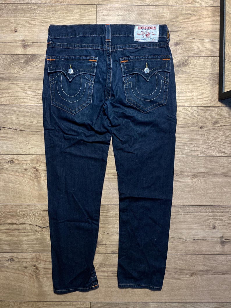 May include: Dark blue denim jeans with white stitching and orange accents. The jeans have two back pockets with a signature curved stitch design and a True Religion brand patch. The jeans are displayed on a wooden surface.