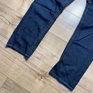 May include: Dark blue denim jeans with visible stitching. The jeans are laid out on a light brown wooden surface. The stitching is a contrasting light tan color. The jeans appear to be new.