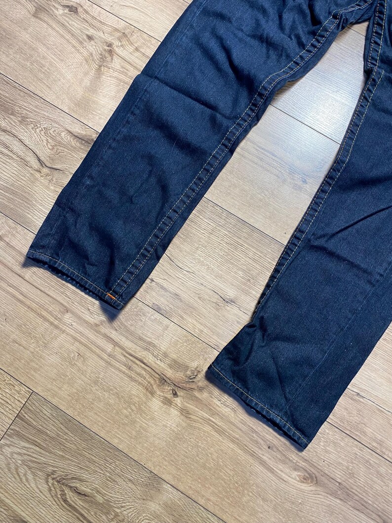 May include: Dark blue denim jeans with visible stitching. The jeans are laid out on a light brown wooden surface. The stitching is a contrasting light tan color. The jeans appear to be new.