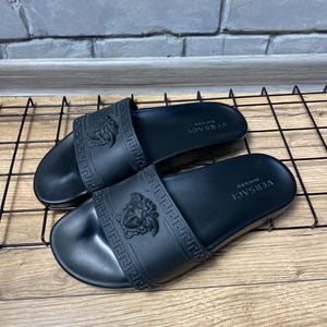 May include: A pair of black rubber slides featuring a wide strap with an embossed Medusa head logo and a Greek key pattern. The footbed is black with the text VERSACE Made in Italy printed in white on the heel area of each sandal.