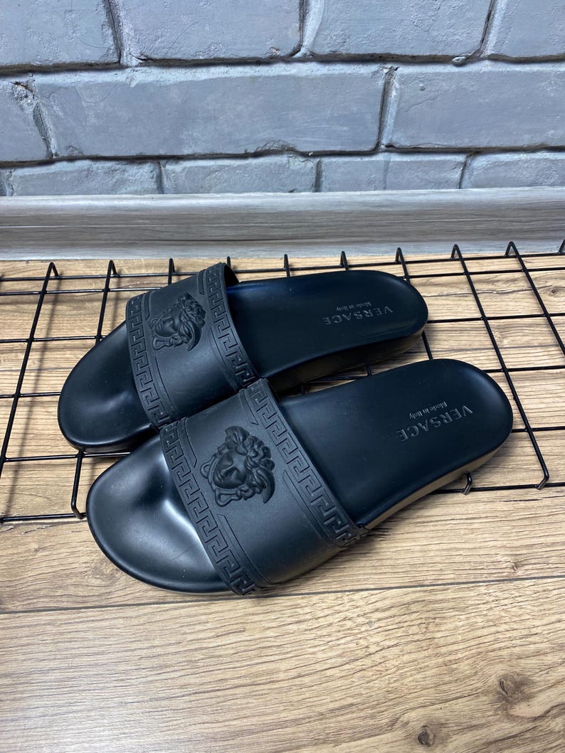 May include: A pair of black rubber slides featuring a wide strap with an embossed Medusa head logo and a Greek key pattern. The footbed is black with the text VERSACE Made in Italy printed in white on the heel area of each sandal.