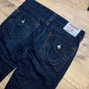 May include: Dark blue denim jeans with white stitching and orange accents. The jeans feature two back pockets with a signature curved stitch design and a True Religion label. The jeans are laid flat on a wooden surface.