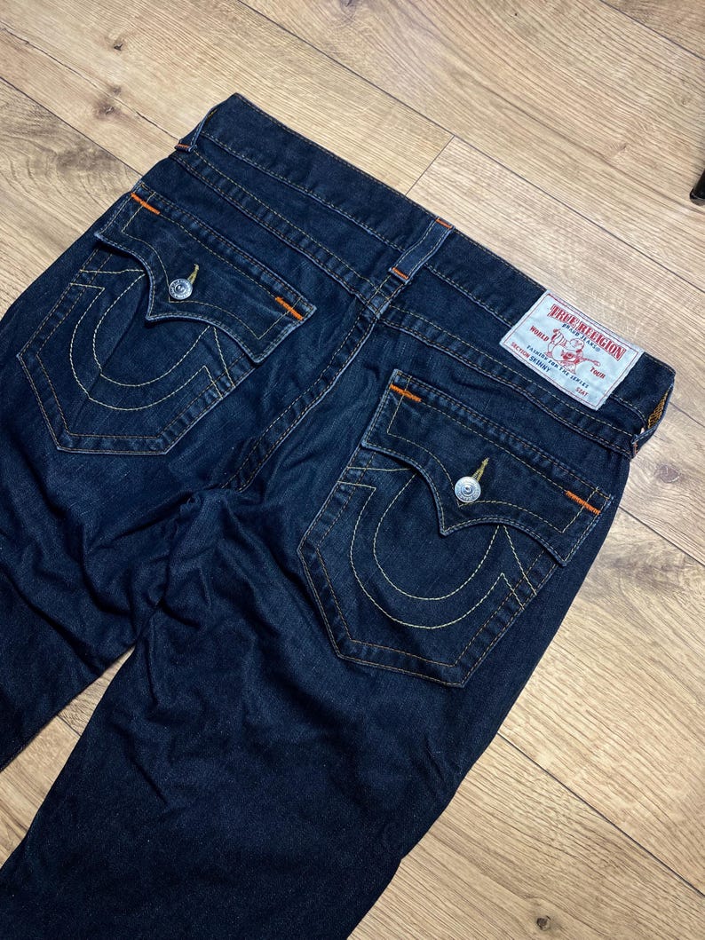 May include: Dark blue denim jeans with white stitching and orange accents. The jeans feature two back pockets with a signature curved stitch design and a True Religion label. The jeans are laid flat on a wooden surface.