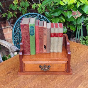 May include: A wooden desktop book rack with a single drawer featuring a decorative metal pull. The rack holds a row of nine vintage hardcover books with various coloured spines, including red, green, and tan, standing upright on the shelf surface.
