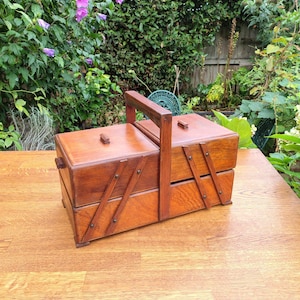 May include: A rectangular wooden sewing box with a medium brown finish, featuring a central vertical handle, two hinged lids with small rectangular knobs, and an accordion-style expandable frame with visible metal rivets on the side panels.