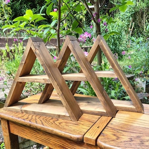 May include: Three wooden A-frame shelves with a rustic, weathered appearance. The shelves are stained a warm brown and feature a single shelf. The shelves are displayed on a wooden table in a garden setting. The shelves are approximately 30 cm wide.