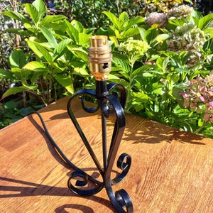 May include: A black metal lamp base with a brass bulb socket. The base has a heart-shaped design with decorative scrollwork. The lamp base is on a wooden surface, with green foliage in the background.