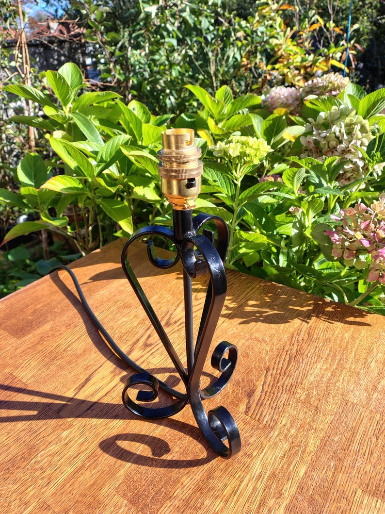 May include: A black metal lamp base with a brass bulb socket. The base has a heart-shaped design with decorative scrollwork. The lamp base is on a wooden surface, with green foliage in the background.
