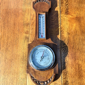 May include: A wooden weather station featuring a thermometer and a barometer. The thermometer shows both Fahrenheit and Celsius scales. The barometer has a silver face with "Rain", "Change", and "Fair" markings. The wood has a warm, reddish-brown hue.