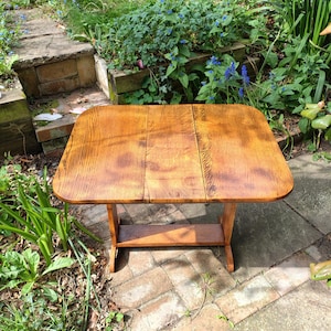 May include: A small, antique wooden table with a rectangular top and a lower shelf. The table is made of light brown wood and has a natural wood grain finish. The table is approximately 76 cm long and 51 cm wide.