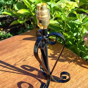 May include: A black metal lamp base with a gold-colored bulb socket. The base has a decorative heart-shaped design and scrollwork. The lamp base is on a wooden surface, with green foliage in the background.