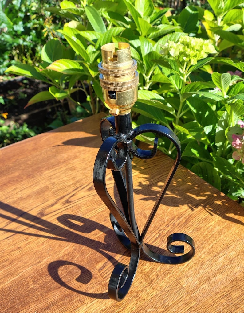 May include: A black metal lamp base with a gold-colored bulb socket. The base has a decorative heart-shaped design and scrollwork. The lamp base is on a wooden surface, with green foliage in the background.