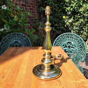 May include: A polished brass table lamp base featuring a smooth, rounded pedestal design with a wide circular base and a central vertical column. The lamp includes a standard light bulb socket at the top and a brown electrical cord coiled at the side.