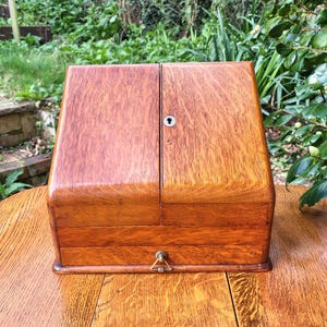 May include: A vintage wooden box in a warm, reddish-brown hue. The box features a hinged lid with a keyhole and a small drawer with a brass pull. It rests on a wooden table, with foliage visible in the background.