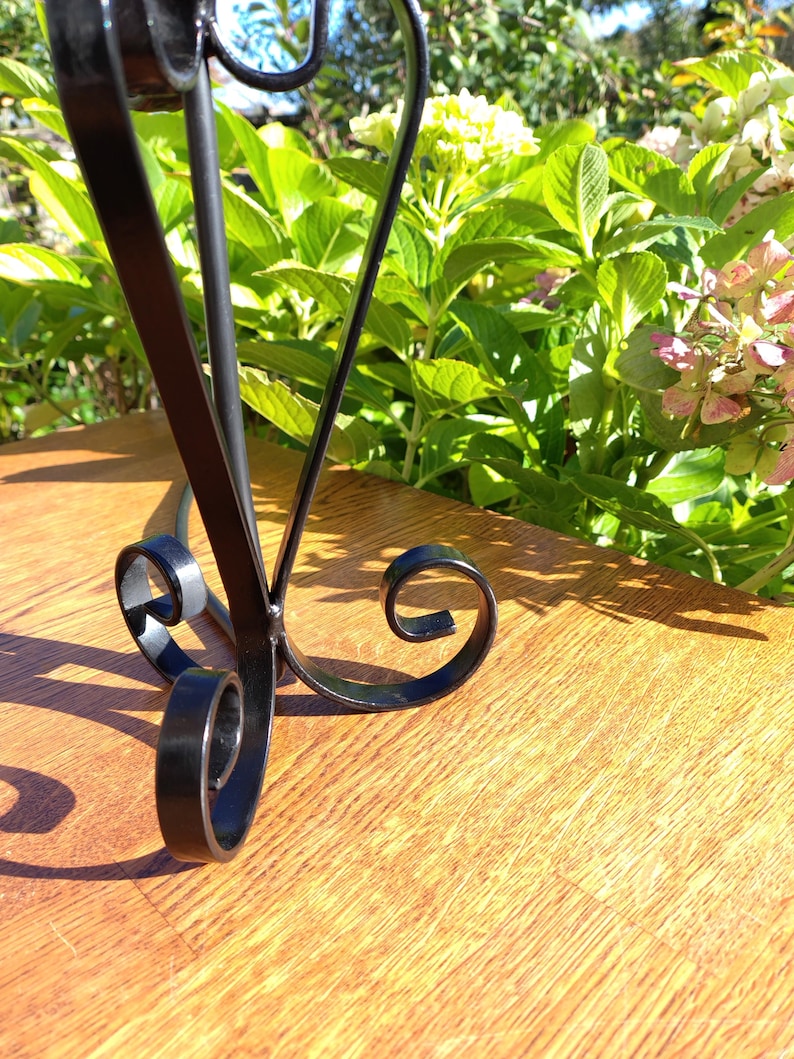 May include: A black metal decorative stand with a scrolled base and curved accents. The stand is on a wooden surface, with green foliage and flowers in the background. The stand is likely for displaying decorative items.
