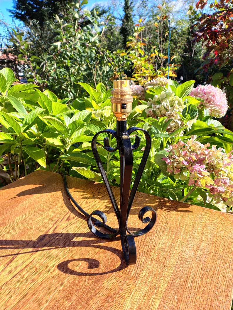 May include: A black metal table lamp base with a heart-shaped design. The lamp base has a gold-colored bulb socket and a black power cord. The lamp base is on a wooden surface, with green foliage and pink flowers in the background.