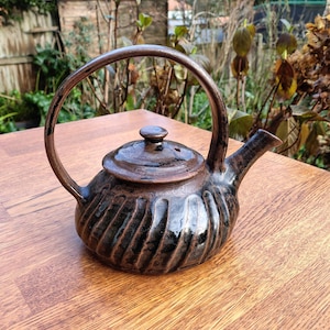 May include: A dark brown ceramic teapot with a curved handle and a lid. The teapot features a ribbed design and a spout. It sits on a wooden surface. The teapot is likely for brewing tea.
