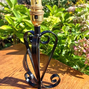 May include: A black metal lamp base with a decorative heart-shaped design. The lamp base has a brass-colored bulb socket. The base is on a wooden surface with green foliage in the background.