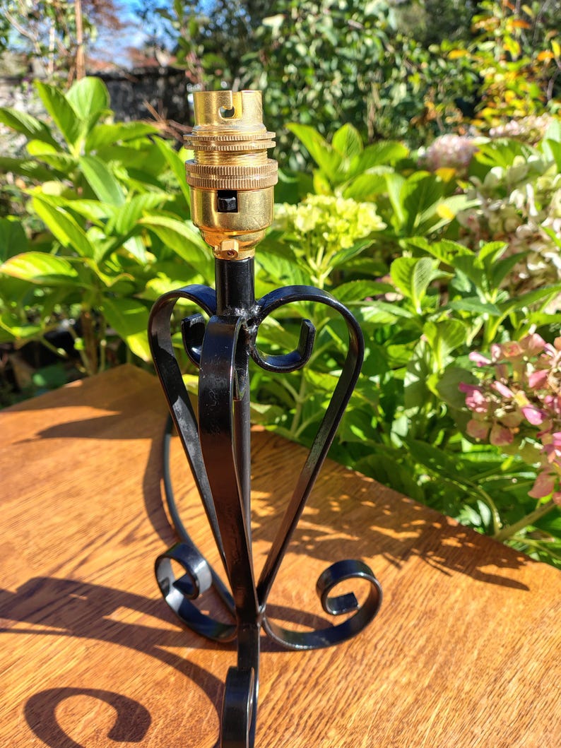 May include: A black metal lamp base with a decorative heart-shaped design. The lamp base has a brass-colored bulb socket. The base is on a wooden surface with green foliage in the background.