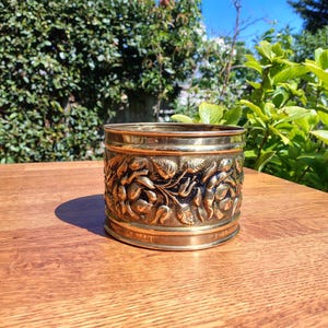 May include: A cylindrical brass planter with an embossed floral design. The planter is made of shiny brass and sits on a wooden surface. The background features green foliage and a bright blue sky.