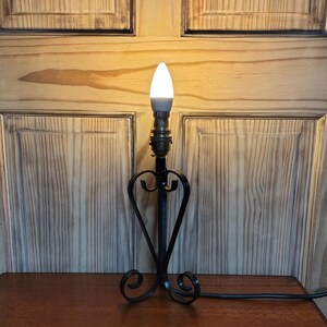 May include: A black wrought iron table lamp with a heart-shaped design. The lamp has a white, teardrop-shaped bulb and a brass-colored socket. The lamp is on a wooden table and has a black power cord.