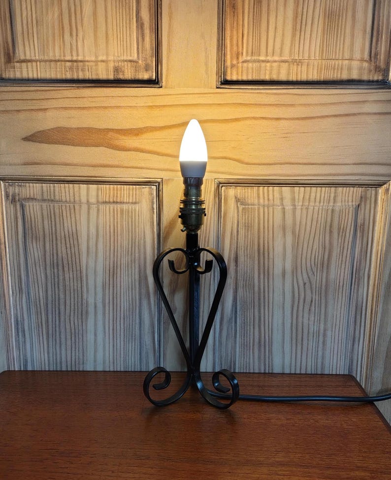 May include: A black wrought iron table lamp with a heart-shaped design. The lamp has a white, teardrop-shaped bulb and a brass-colored socket. The lamp is on a wooden table and has a black power cord.