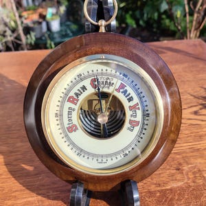 May include: A classic wooden barometer with a circular dial. The dial indicates weather conditions with labels like "Stormy" and "Fair." The barometer has a dark wood finish, a brass ring, and stands on small black feet. The dial is measured in millibars.