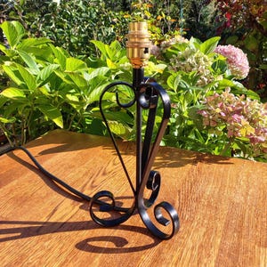 May include: Black wrought iron table lamp with a heart-shaped design. The lamp has a brass bulb socket and a decorative base with scrolled accents. The lamp is on a wooden surface, with greenery in the background.